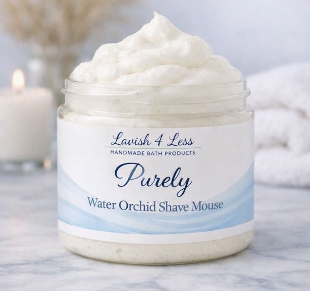 Jar of Purely Water Orchid Shave Mousse by Lavish 4 Less Handmade Bath Products, filled with creamy, whipped mousse on a white marble surface. The label features soft blue watercolor accents and spa‑style typography. A lit candle, rolled towel, and dried pampas grass create a calm, luxurious background.