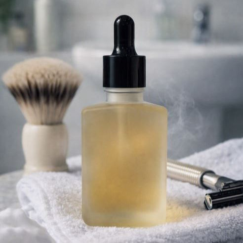 Frosted glass dropper bottle of shave oil resting on a folded white towel beside a shaving brush and metal razor, with soft bathroom lighting and a blurred sink background.