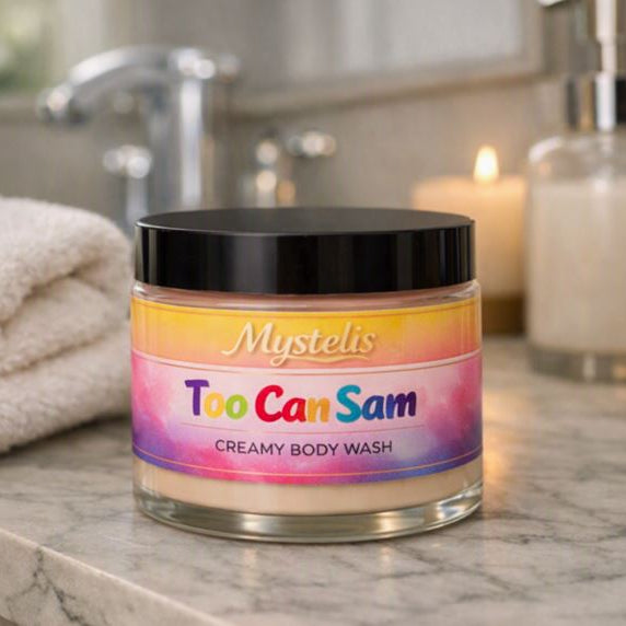 Clear jar of Mystelis Too Can Sam Creamy Body Wash with a black lid, placed on a marble bathroom counter beside a folded white towel, a white soap dispenser, and a softly glowing candle, with a blurred faucet and mirror in the background.