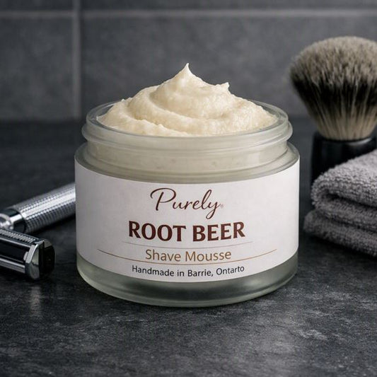 Purely Root Beer Shave Mousse — open frosted jar on dark slate bathroom counter with chrome razor, black shaving brush, and gray towel, cool masculine lighting, handmade in Barrie Ontario.