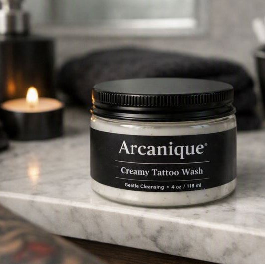 4 oz clear plastic jar with black lid labeled “Arcanique Creamy Tattoo Wash” placed in a tattoo‑lover bathroom scene with candlelight, black towel, soap dispenser, and accessories on a stone countertop.