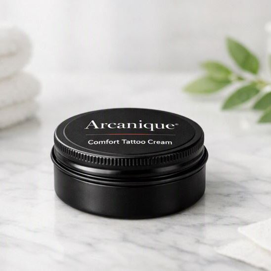 Small black screw‑lid tin labeled “Arcanique Comfort Tattoo Cream” placed on a bright white marble spa‑studio surface with soft daylight, folded towels, and greenery in the background.