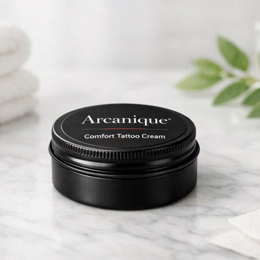 Small black screw‑lid tin labeled “Arcanique Comfort Tattoo Cream” placed on a bright white marble spa‑studio surface with soft daylight, folded towels, and greenery in the background.