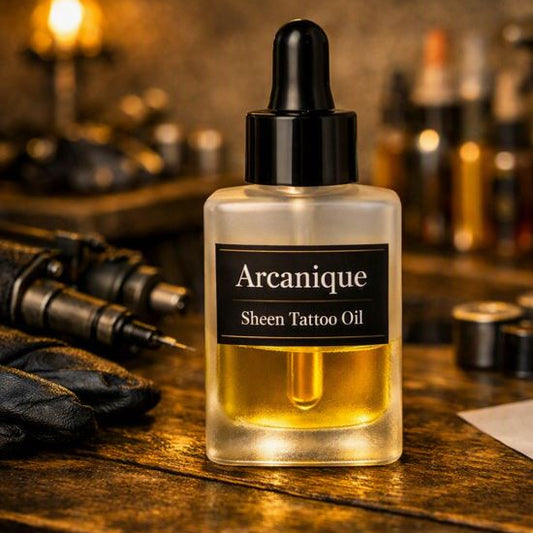 Rectangular 28 ml frosted glass dropper bottle labeled “Arcanique Sheen Tattoo Oil,” half‑filled with golden oil, placed on a tattoo‑studio work surface with blurred inks, gloves, and tattoo machine under warm golden lighting.