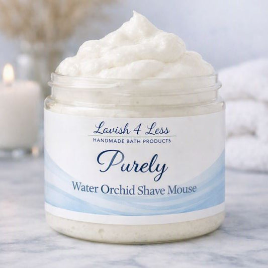 Jar of Purely Water Orchid Shave Mousse by Lavish 4 Less Handmade Bath Products, filled with creamy, whipped mousse on a white marble surface. The label features soft blue watercolor accents and spa‑style typography. A lit candle, rolled towel, and dried pampas grass create a calm, luxurious background.