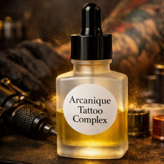 Frosted glass dropper bottle labeled “Arcanique Tattoo Complex,” half‑filled with golden oil, placed on a tattoo‑studio work surface with blurred inks, machine, and warm lighting in the background.