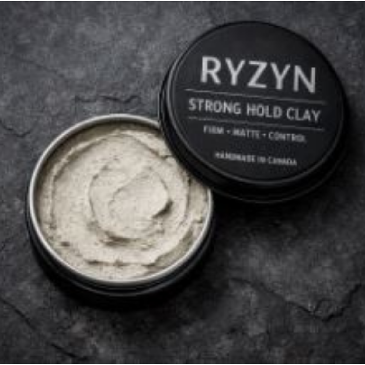 A grooming flatlay featuring an open tin of Ryzyn Strong Hold Clay with its matte black lid showing bold white lettering, placed on a dark textured stone background. The tin reveals dense off‑white clay with a firm, swirled texture, symbolizing strong hold and matte control.