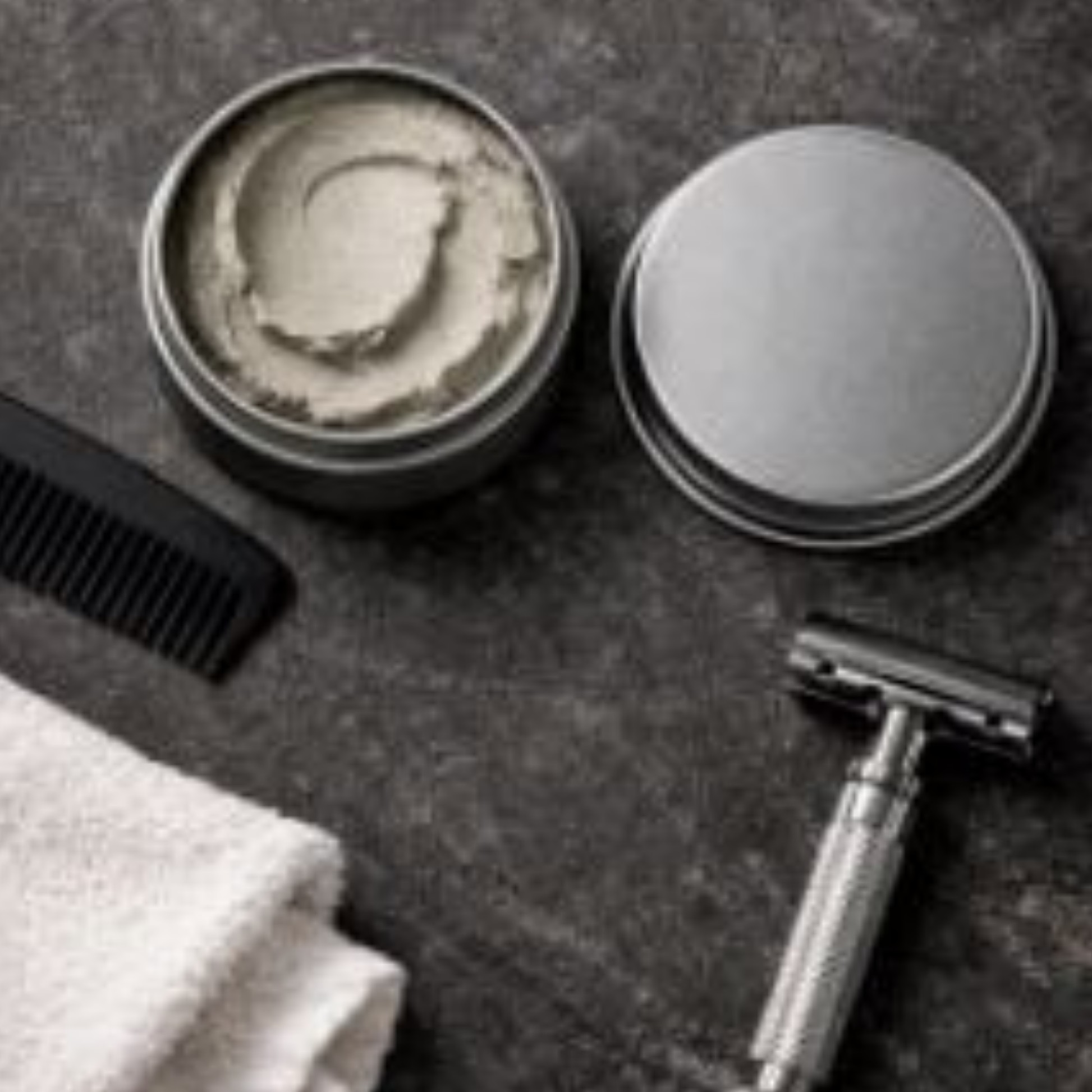 A grooming flatlay featuring an open 2 oz matte hair clay tin with a smooth swirled texture, placed beside a black comb, silver safety razor, and white towel on a dark textured stone background.